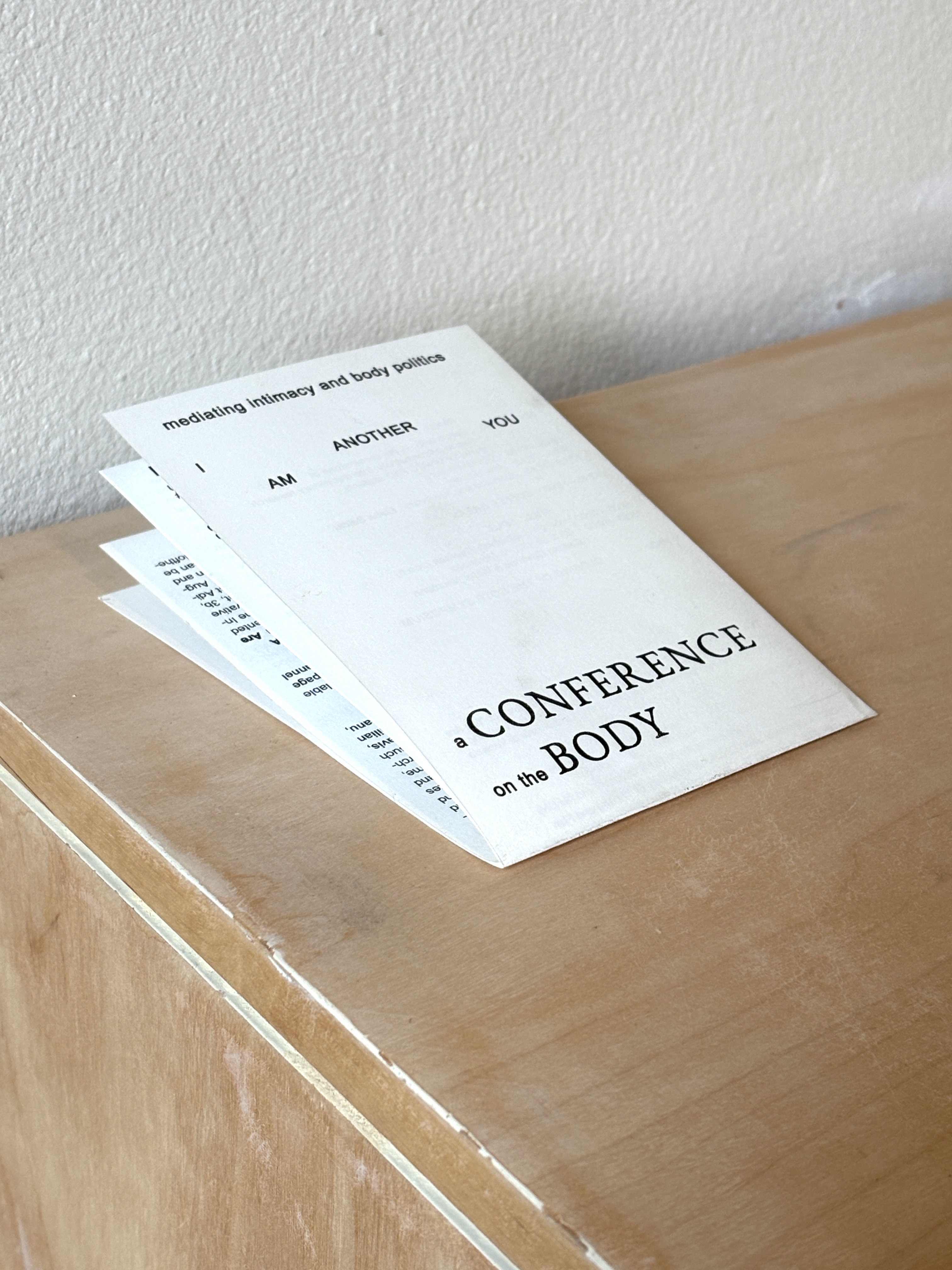 A folded paper titled 'a CONFERENCE on the BODY' on a wooden surface near a white textured wall.
