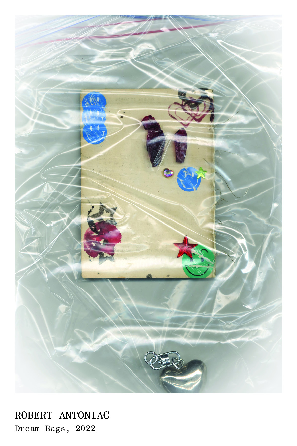 Small beige square paper decorated with stickers and dried flower petals, enclosed in a transparent plastic bag with a silver heart-shaped charm attached.
