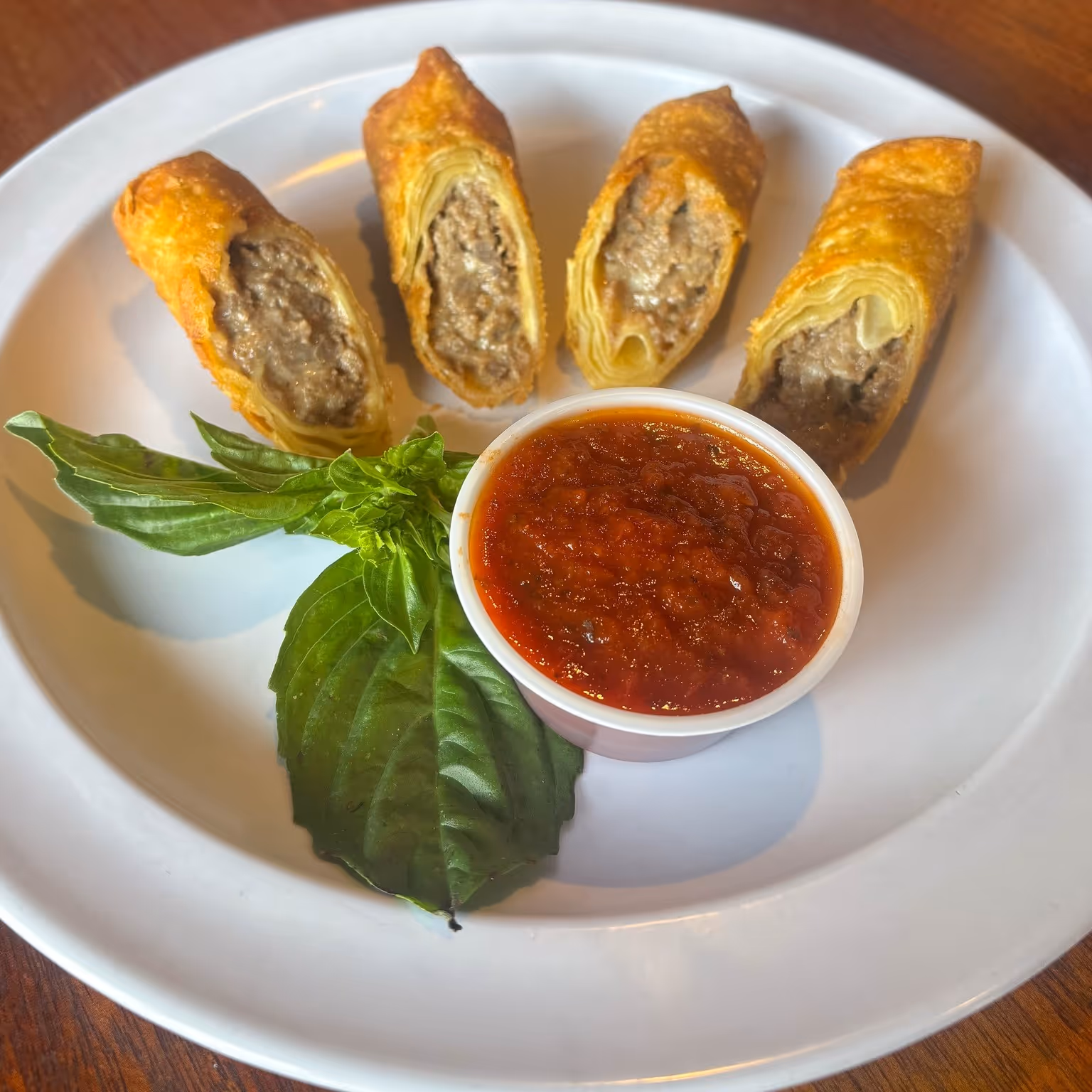 Golden-brown egg rolls filled with savory meat with a cup of rich red marinara sauce and fresh basil leaves