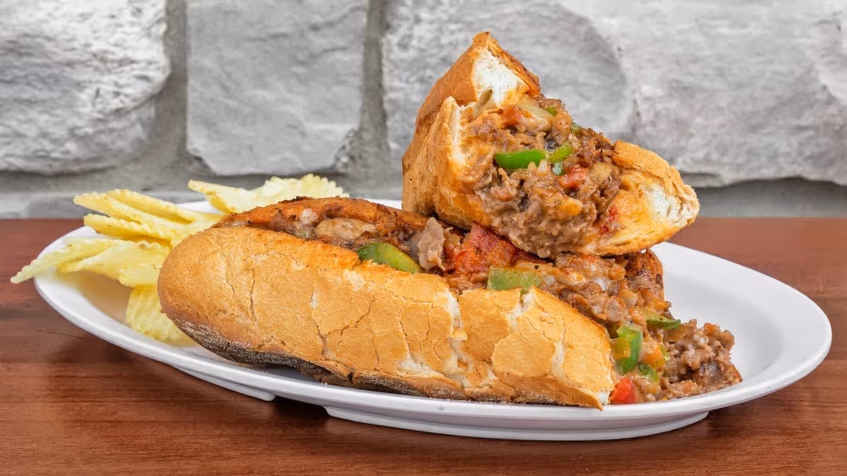 sub filled with ground meat, green peppers, and saucy ingredients sits on a white plate, accompanied by wavy potato chips.