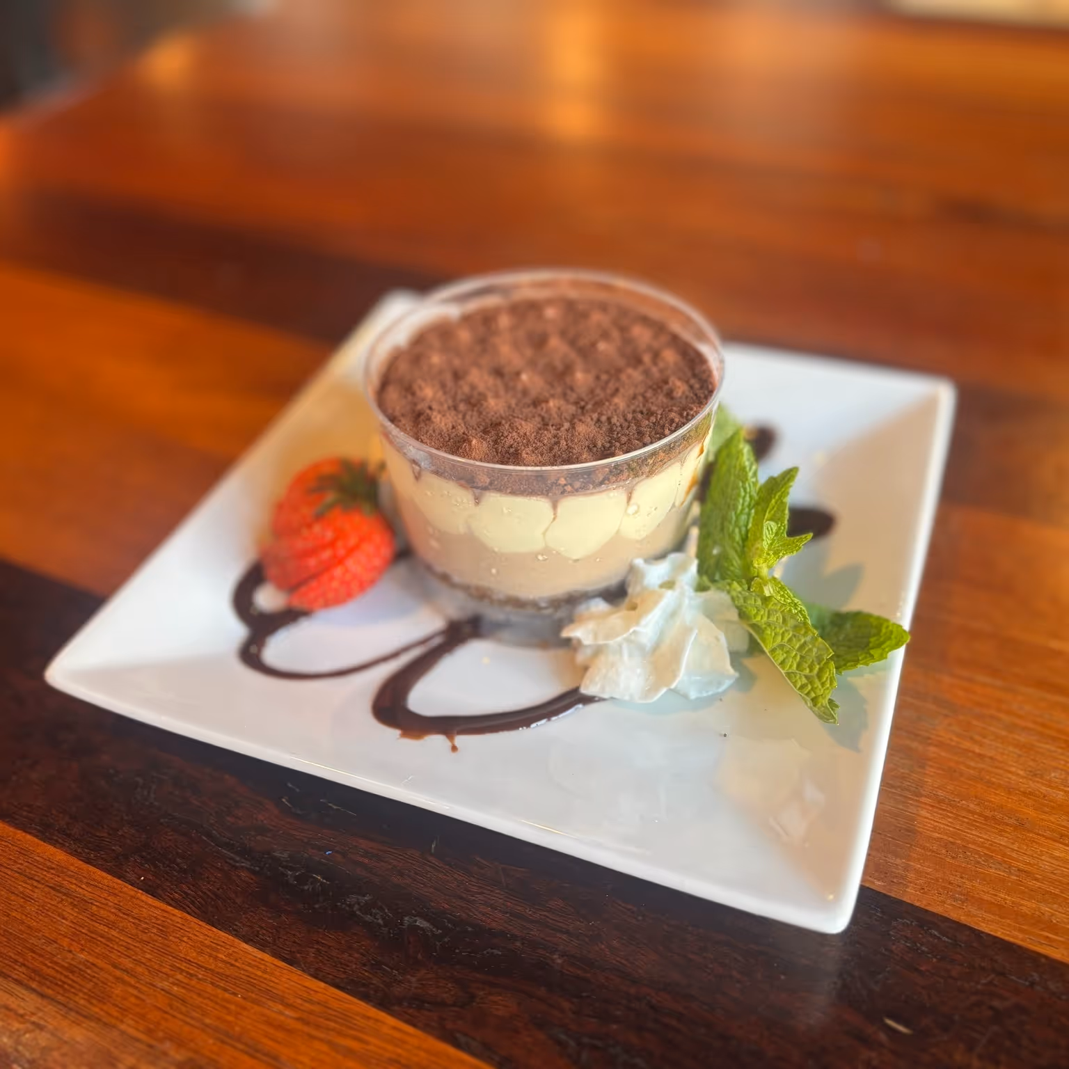 A delectable layered dessert topped with cocoa powder garnished with fresh mint, a strawberry, whipped cream, and chocolate sauce.