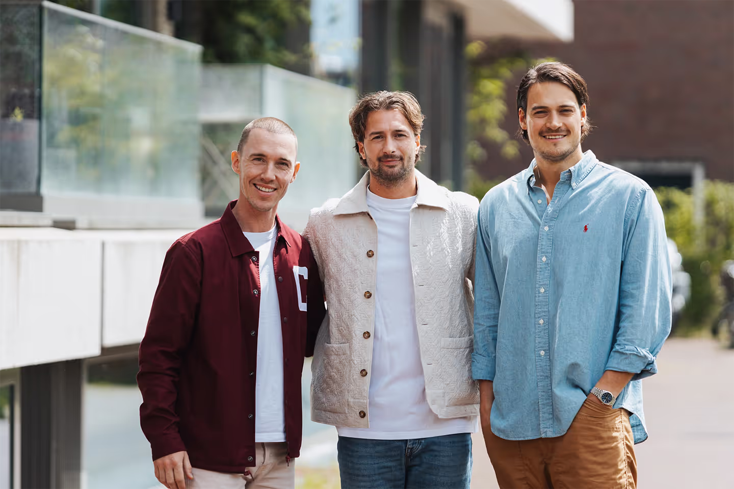 Three men standing outdoors, smiling at the camera, dressed in casual jackets and shirts.