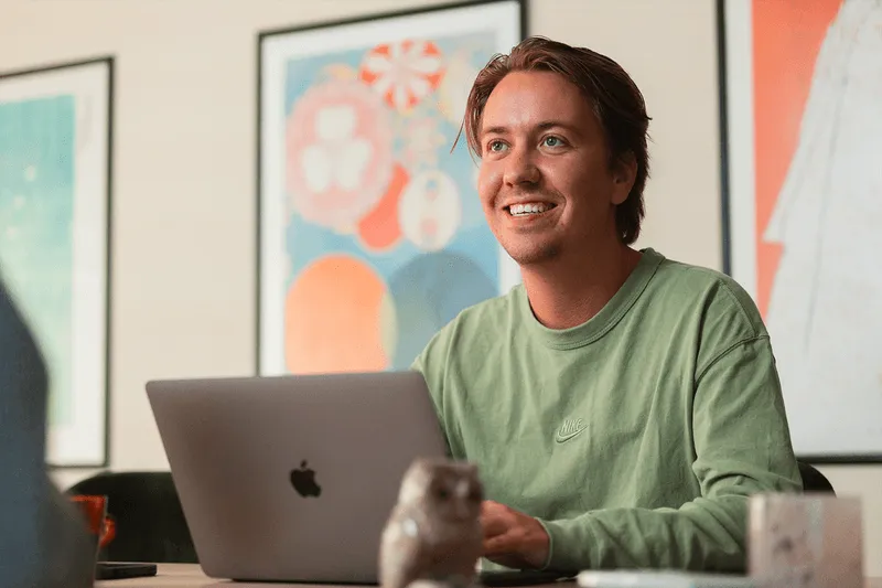 Smiling young man wearing a green Nike sweatshirt working on a laptop at a table with framed artwork on the wall behind.