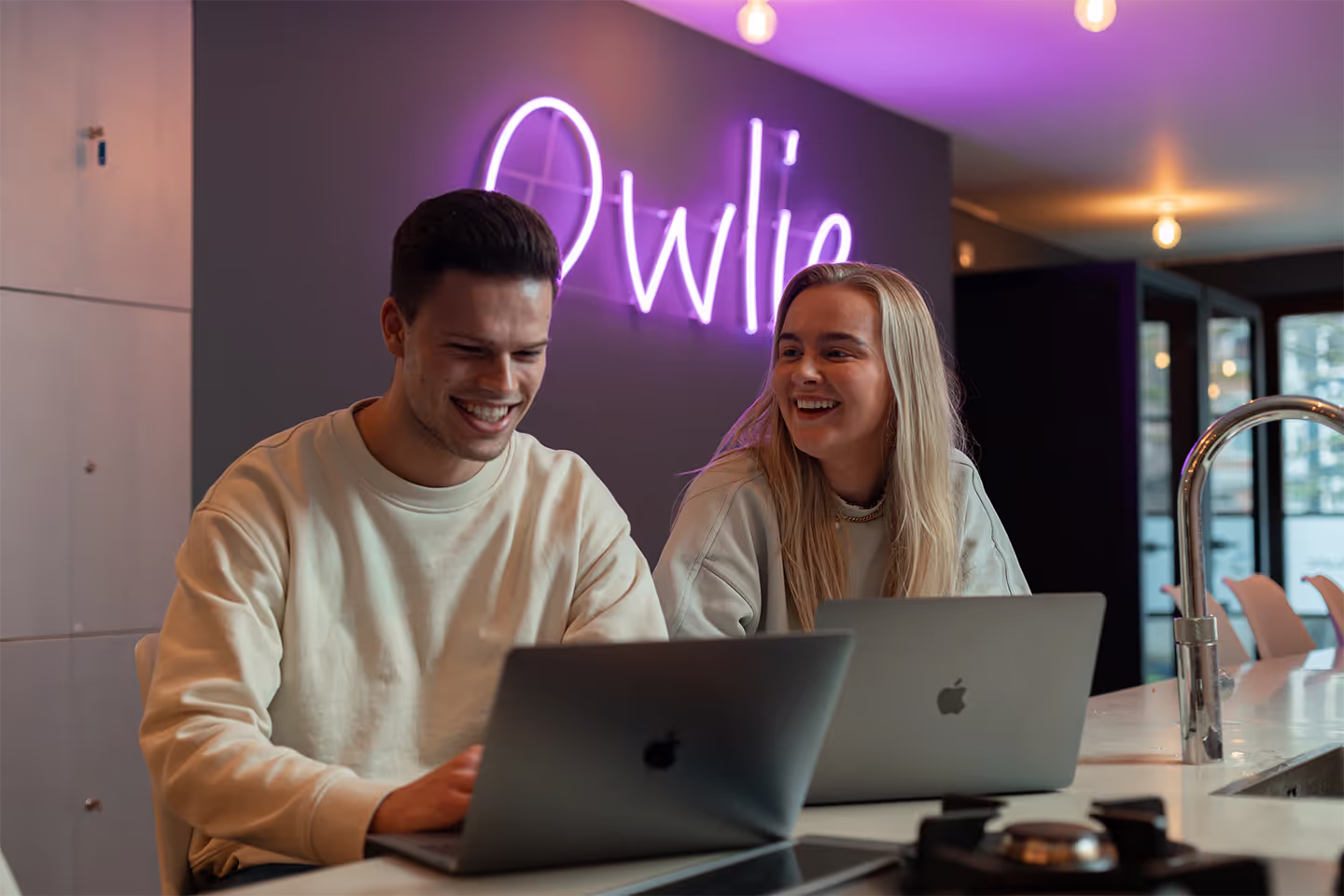 Two young people smiling and working on laptops at a modern kitchen island with a purple neon sign in the background.