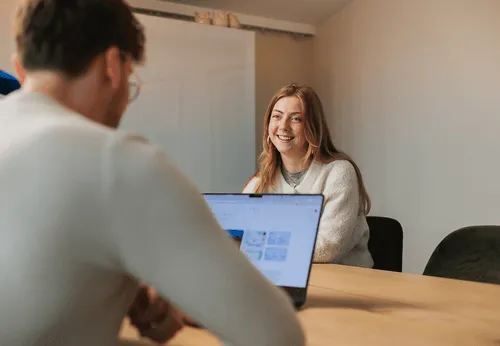 Woman smiling and talking to a man who is working on a laptop at a wooden table in a bright room.