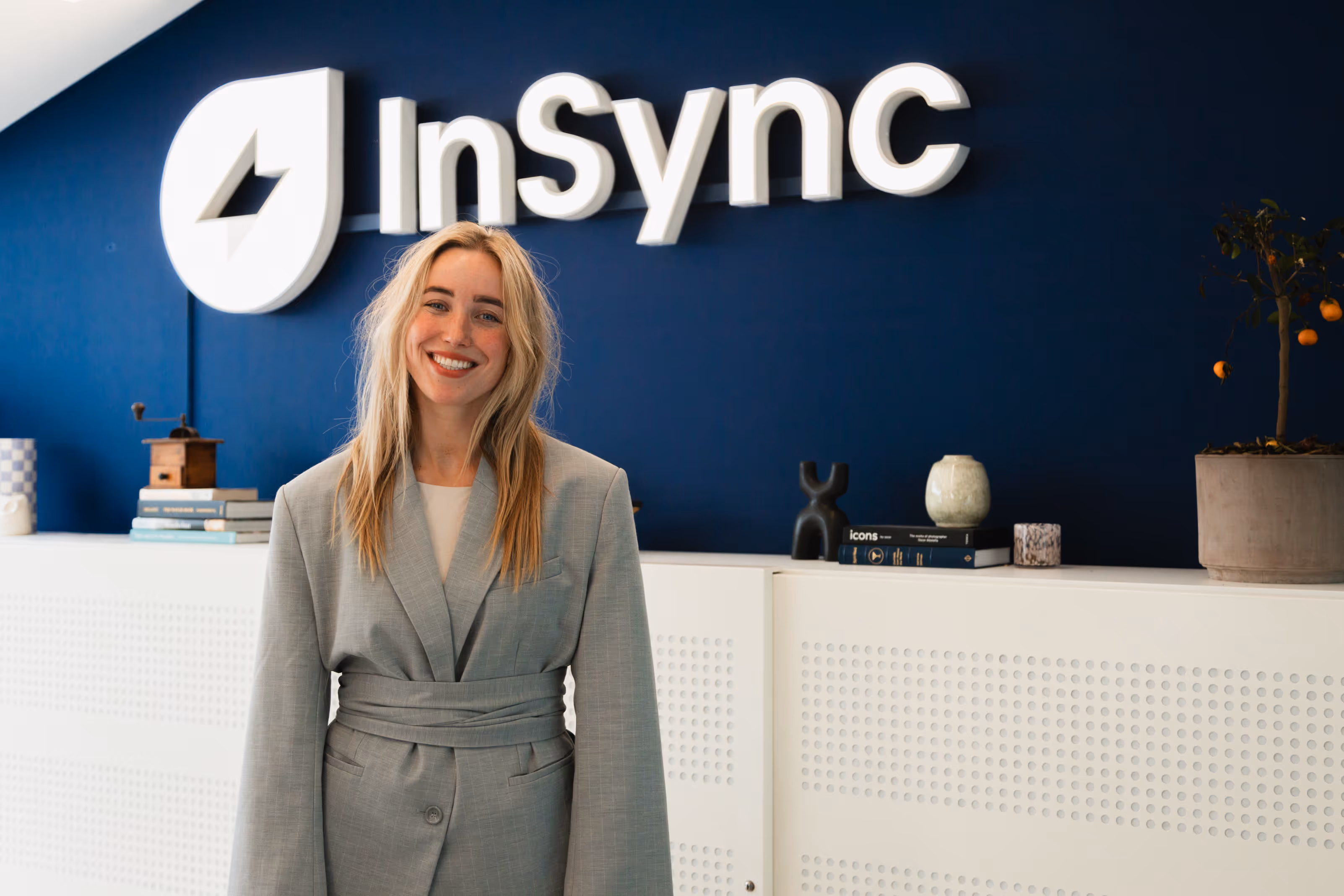 Smiling woman in gray blazer standing in front of a blue wall with the InSync logo and a white cabinet with books and a potted plant.