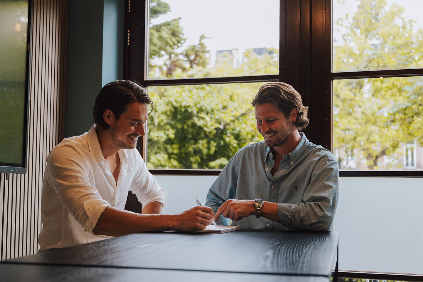 Two men smiling and discussing while one writes on a notepad at a table near a large window with trees outside.