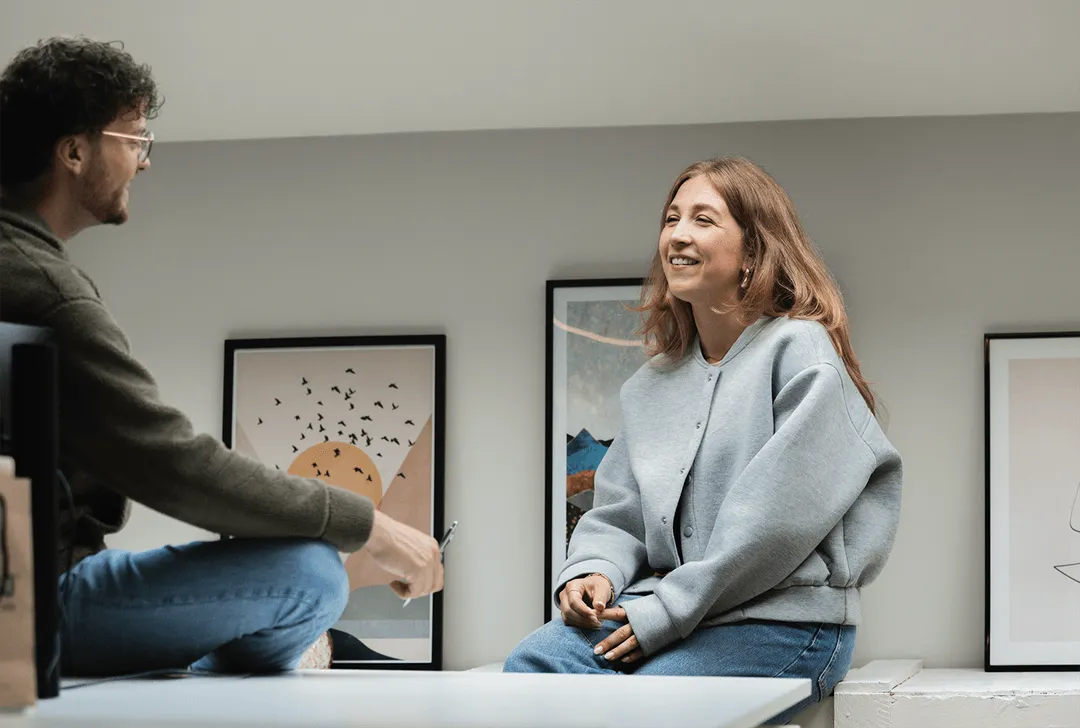 Two people sitting and smiling while having a conversation in a room with framed artwork on the wall.