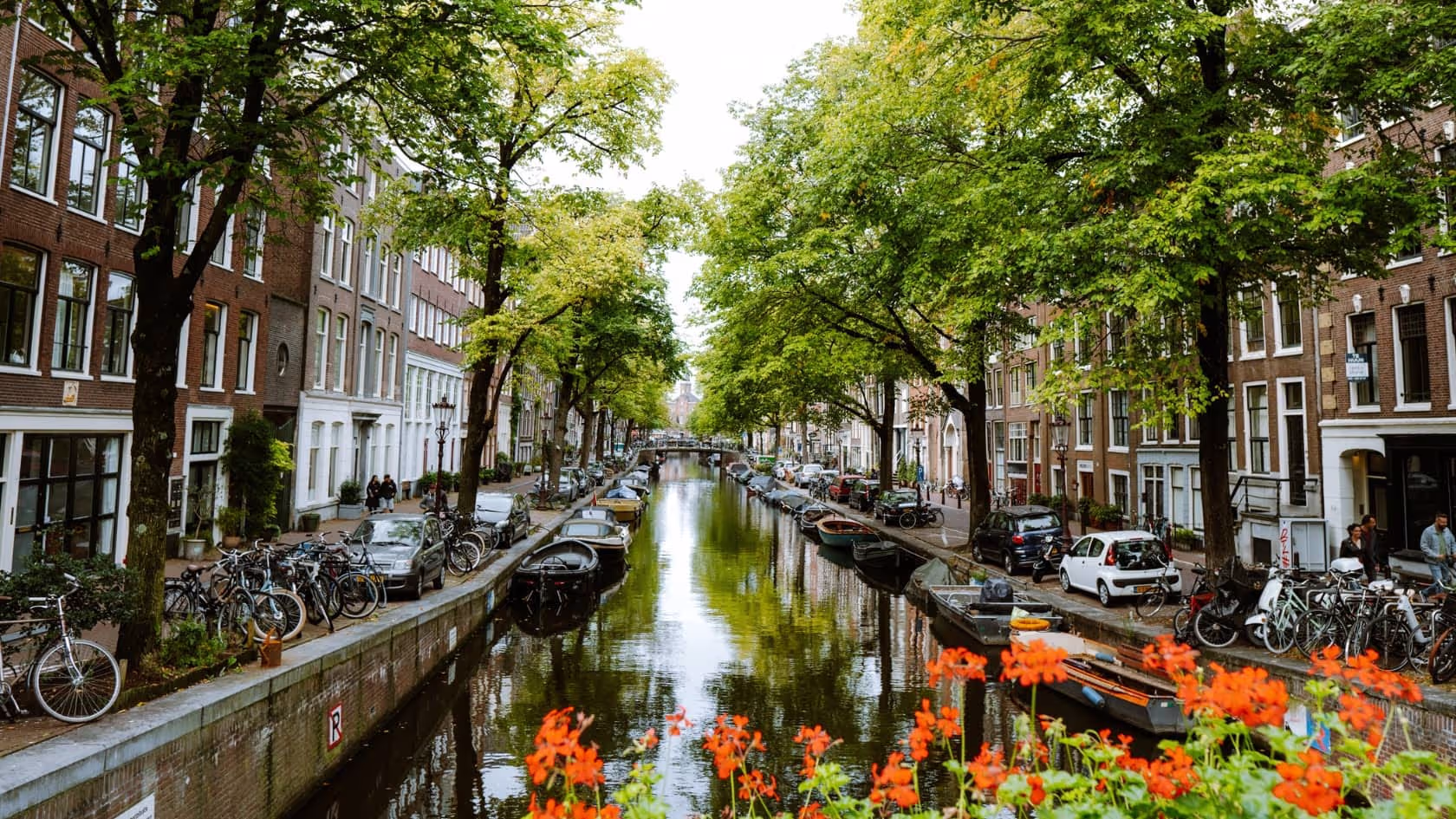 Scenic canal in a city lined with trees, boats, bicycles, and brick buildings with orange flowers in the foreground.