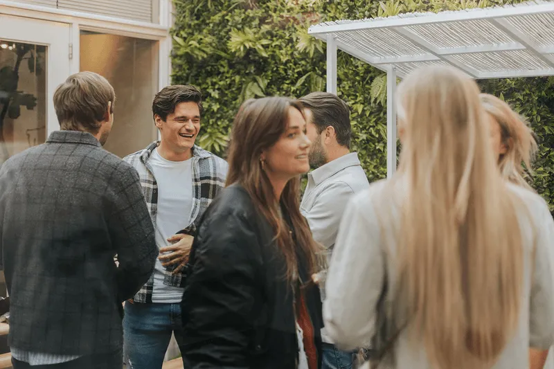 Group of six young adults socializing outdoors near a green wall, smiling and talking.