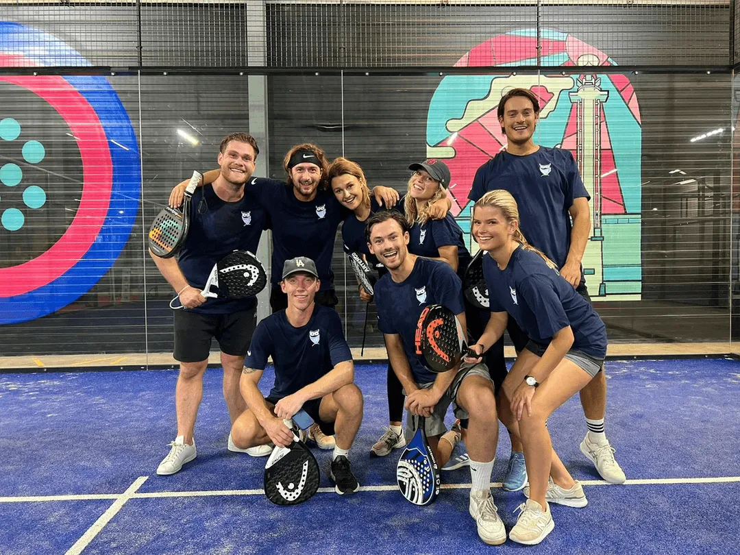 Group of eight smiling young adults in blue sports shirts posing on a blue paddle tennis court holding paddles.