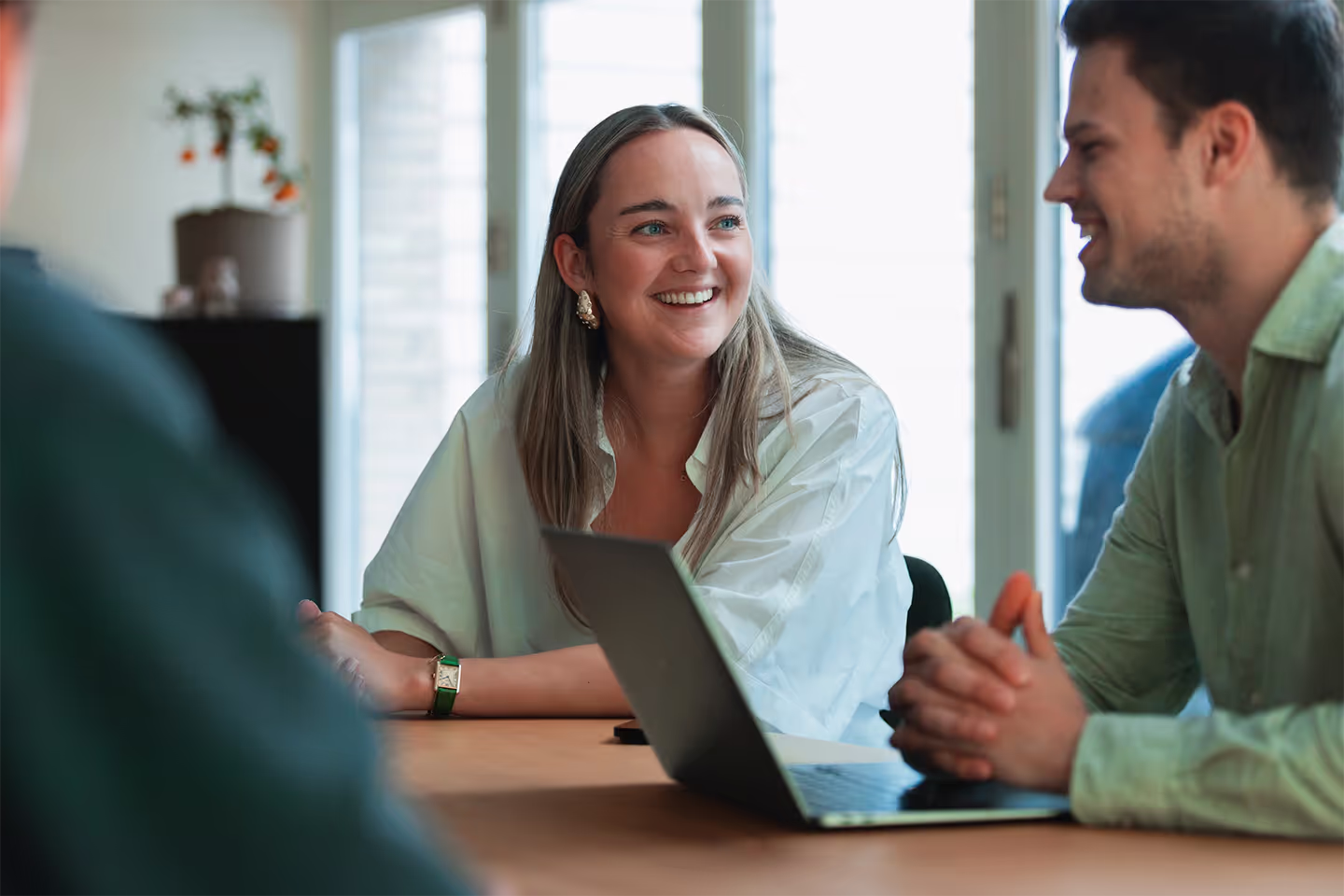 Smiling woman in white shirt talking with two colleagues at a meeting table with a laptop.