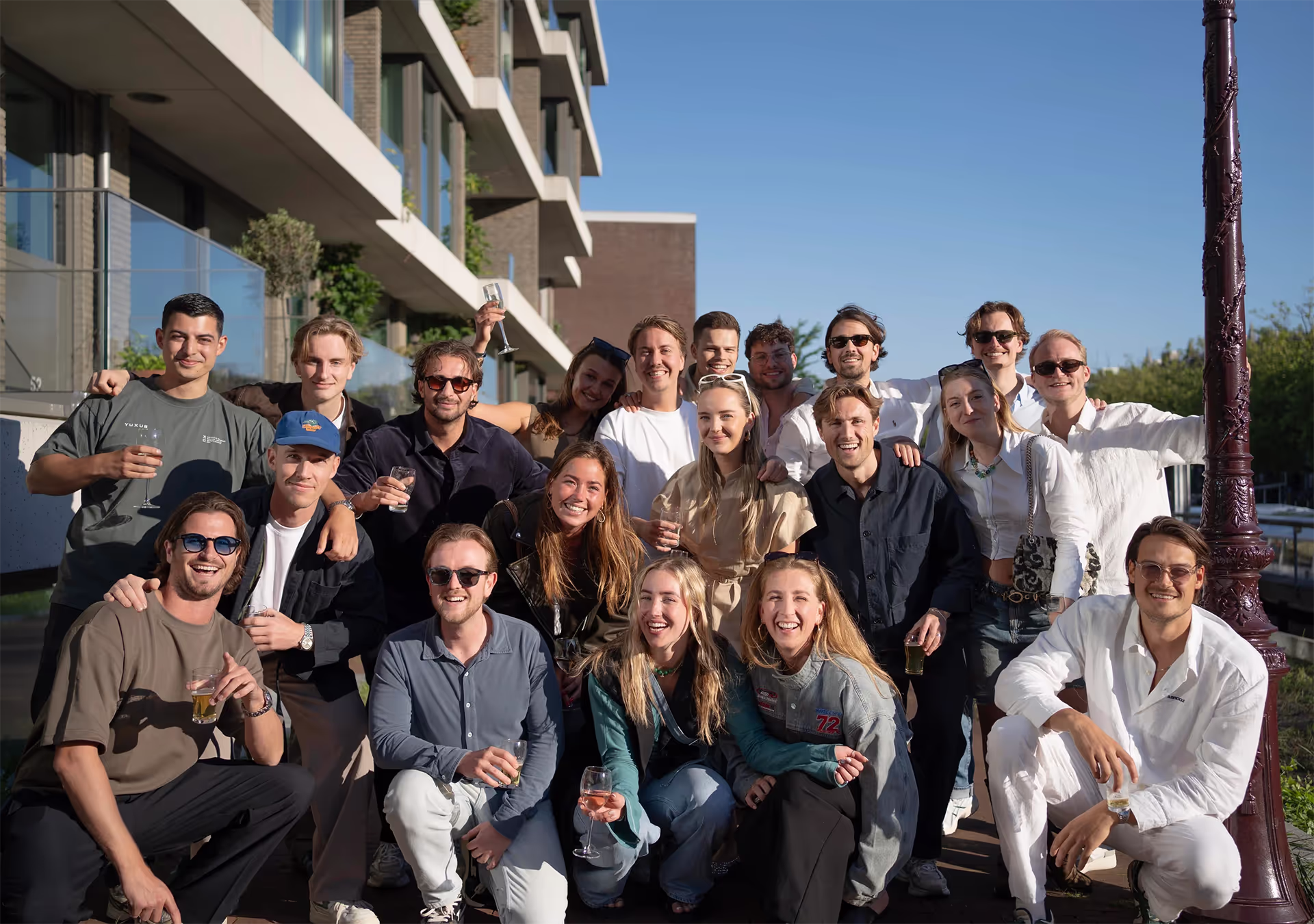 Group of young adults smiling and holding drinks outdoors in front of a modern building under clear blue sky.