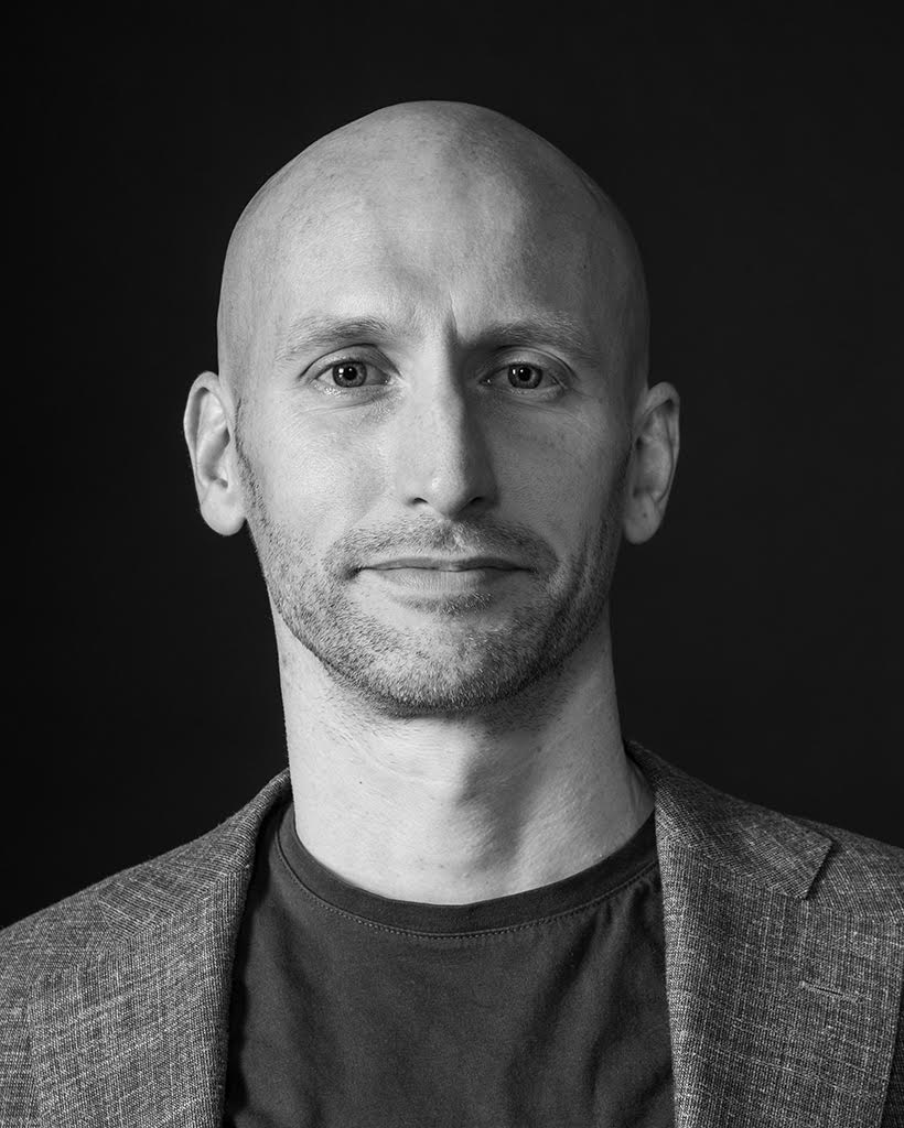 Black and white close-up portrait of Cahl with light facial stubble wearing a dark shirt and a textured blazer against a plain dark background.