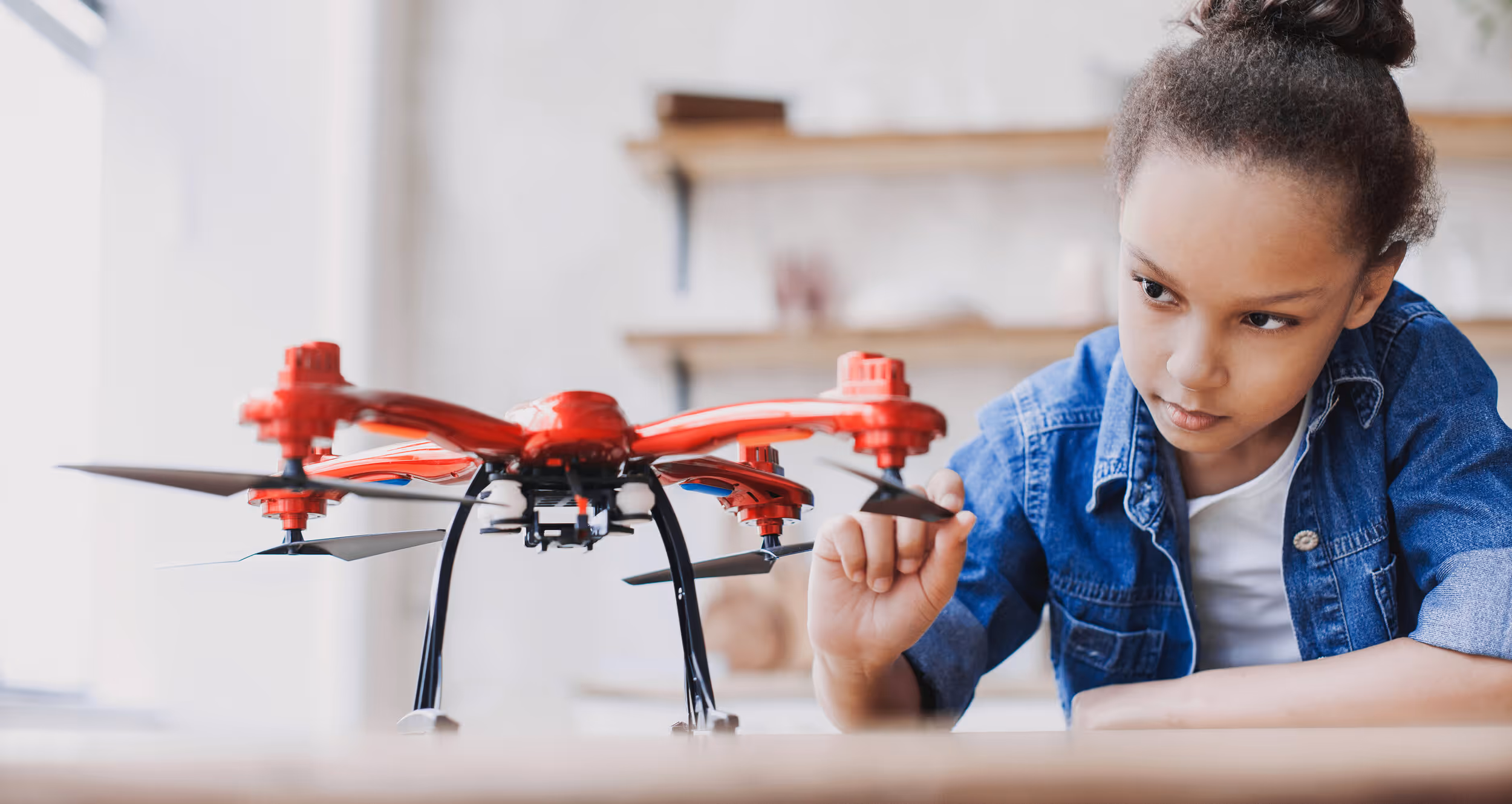 Young student in denim jacket examining red and black drone on table
