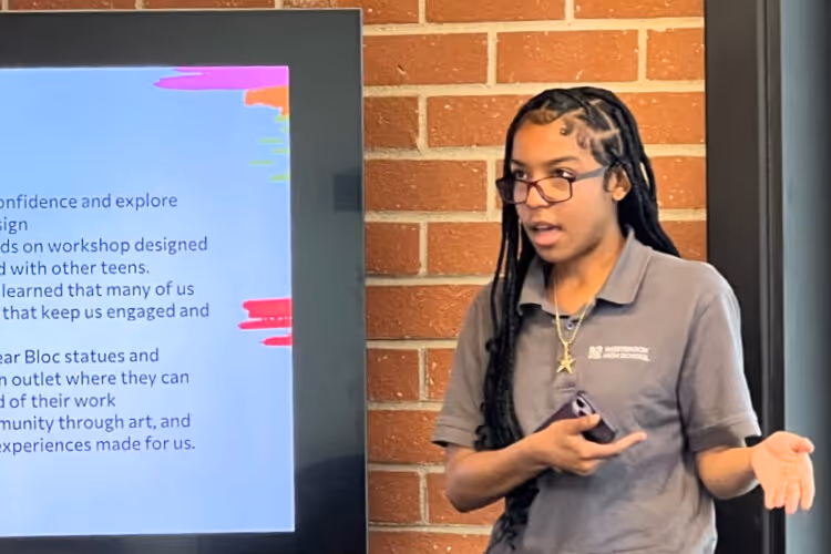 Student presenting at brick wall with text displayed on screen behind her.