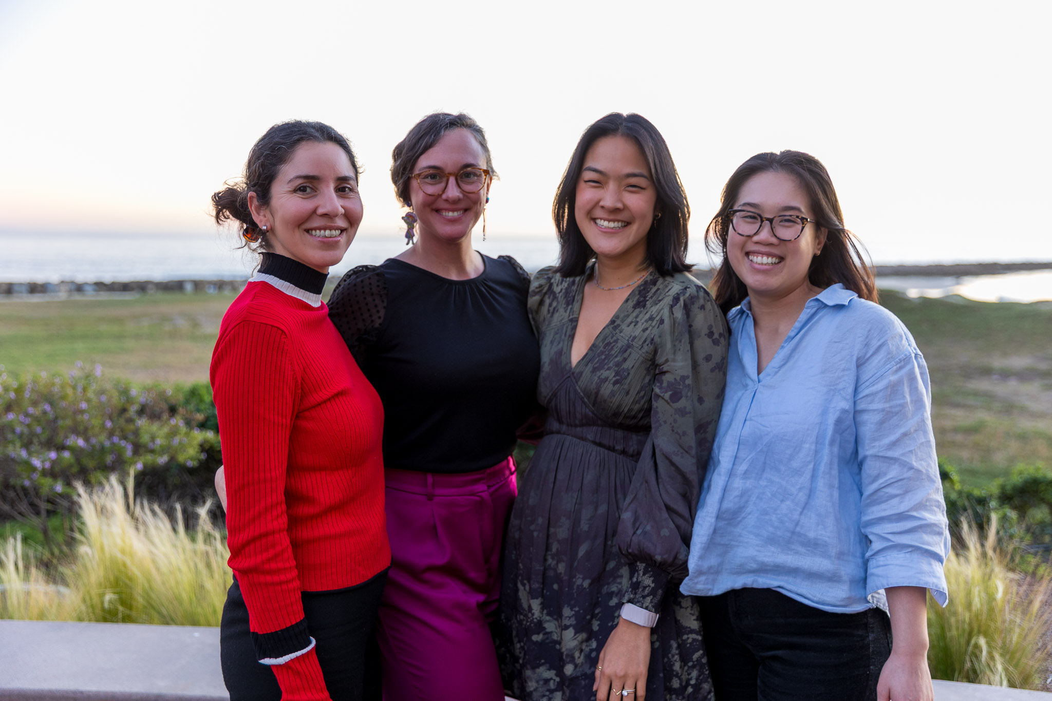 Four smiling colleagues standing together in a scenic coastal landscape at sunset.