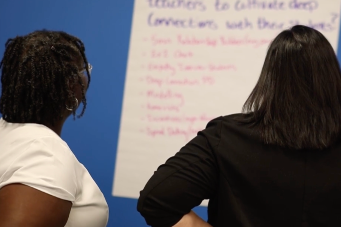 Two women viewing a whiteboard with handwritten text against a blue background.