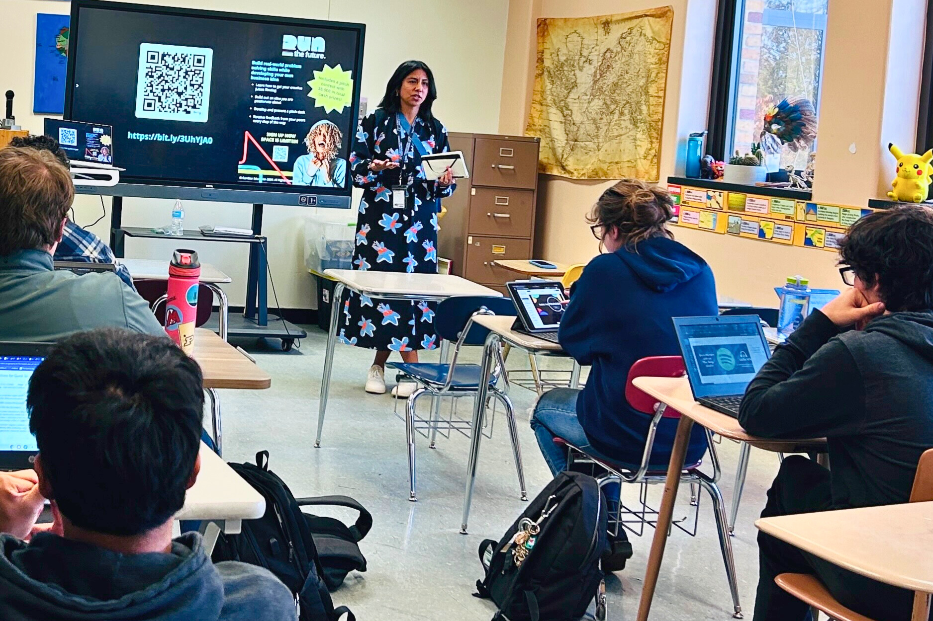 Teacher presenting to students with laptops in classroom with projector display.