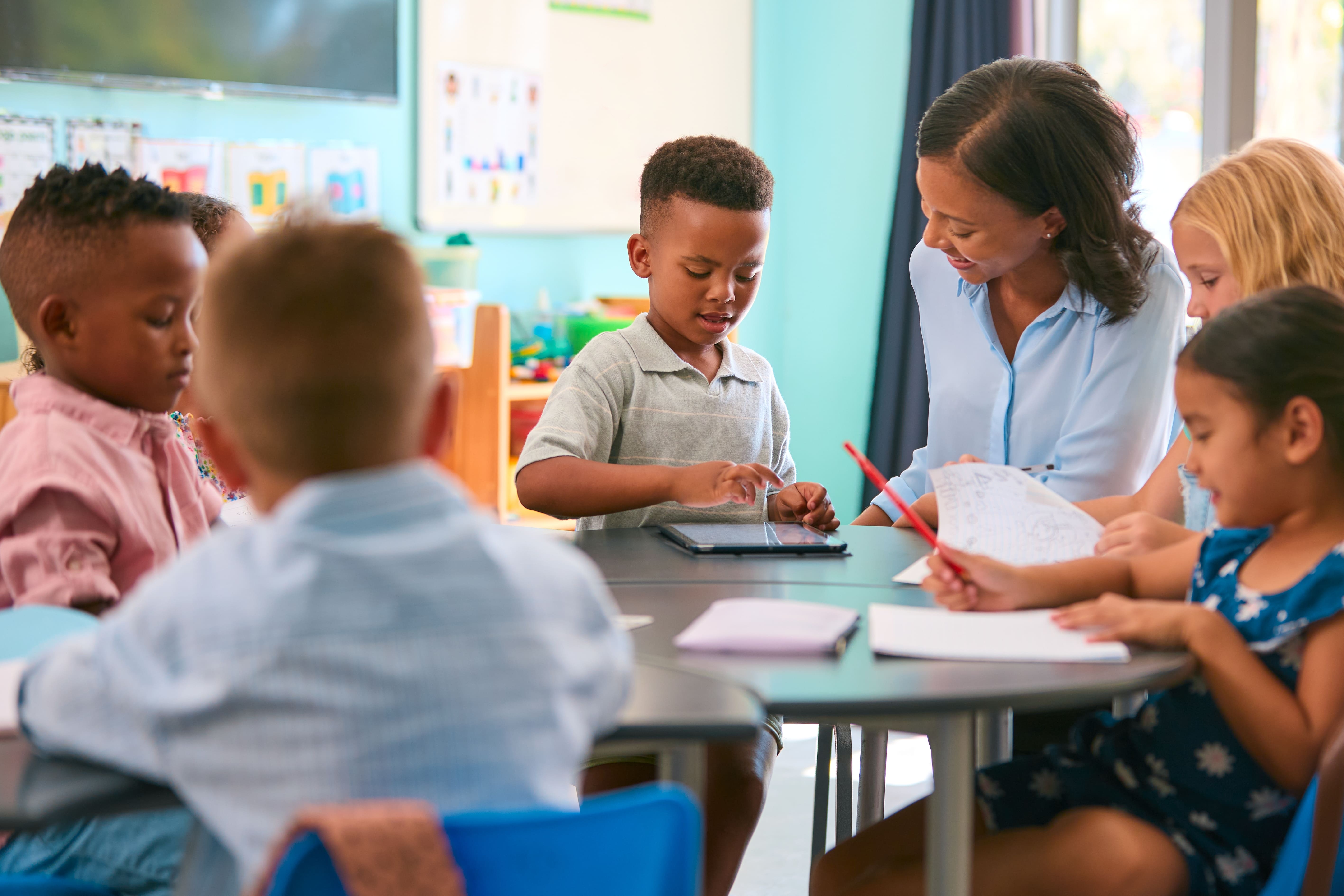 Teacher helping elementary school children with drawing and writing at classroom table