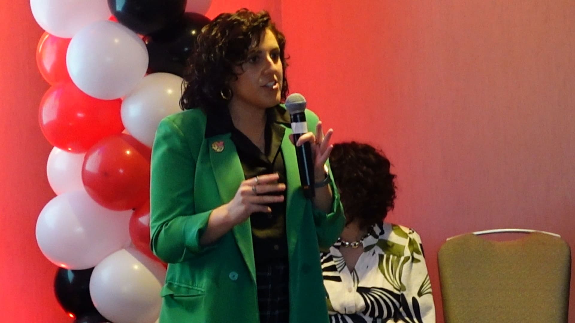 Leader speaking at microphone in green blazer with red and white balloons background.