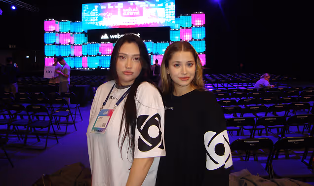 Two young women wearing black and white shirts with matching sleeve logos posing in front of a colorful illuminated backdrop at an event.