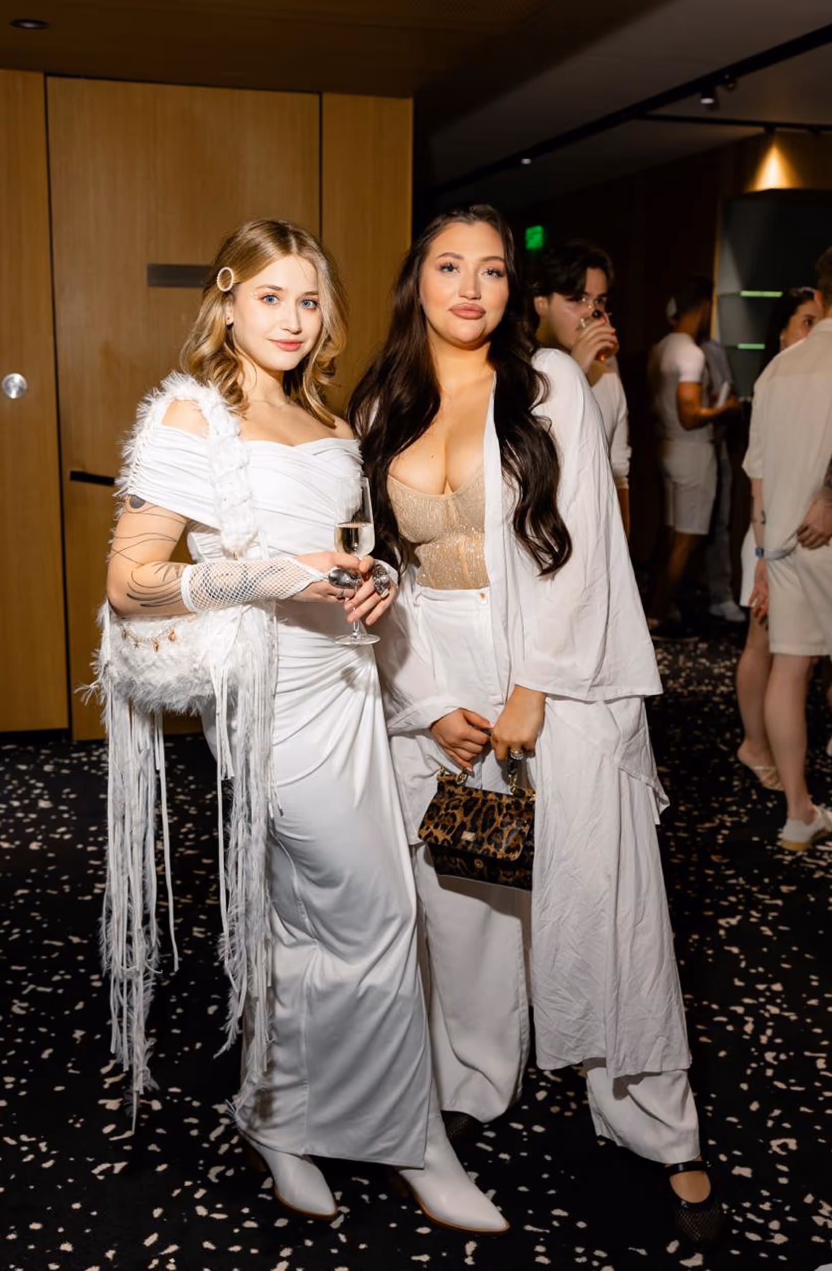 Two women posing at an indoor event, one wearing a white dress, the other in white pants and a sheer beige top holding a leopard print bag.