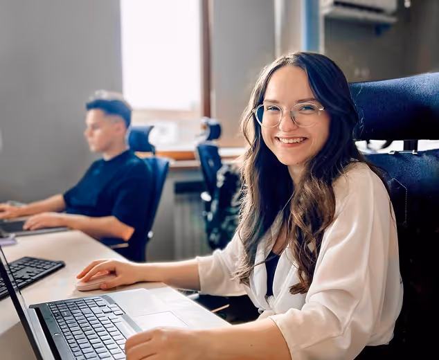 Smiling woman with glasses working on a laptop in an office, with a man blurred in the background.