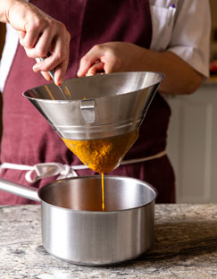 Chef straining Rossafina tomato sauce through a metal sieve into a pot on a kitchen counter.