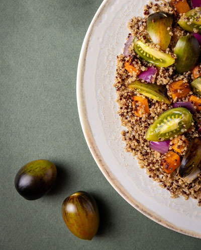 Plate of quinoa salad with roasted vegetables and sliced Midnight plum tomatoes on a green surface with whole Midnight plum tomatoes beside the plate.
