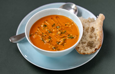 Bowl of creamy tomato and lentil soup garnished with herbs and drizzle, served with a slice of crusty bread on a blue plate.