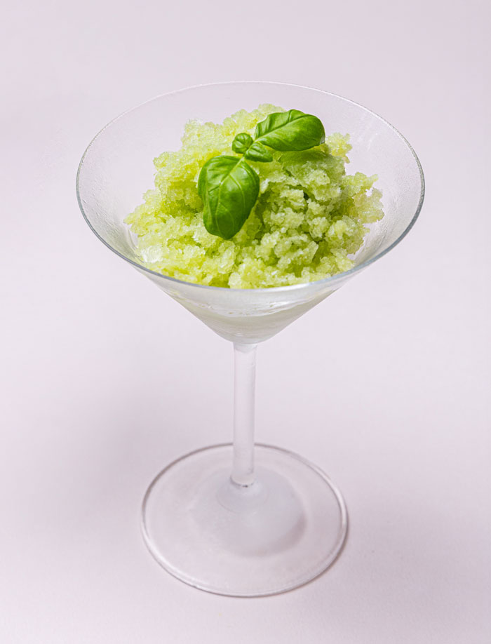 Green granita dessert served in a clear martini glass garnished with a basil leaf.