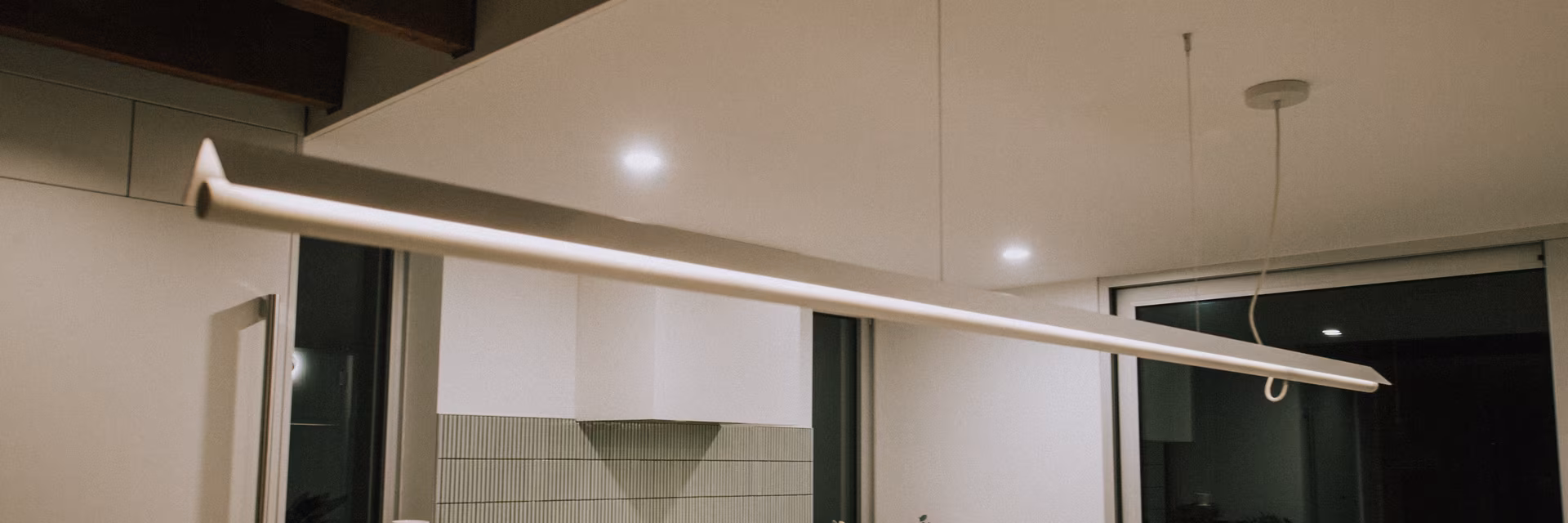 Une lampe linéaire suspendue et allumée dans une cuisine moderne avec des armoires blanches et un mur à carreaux beige.