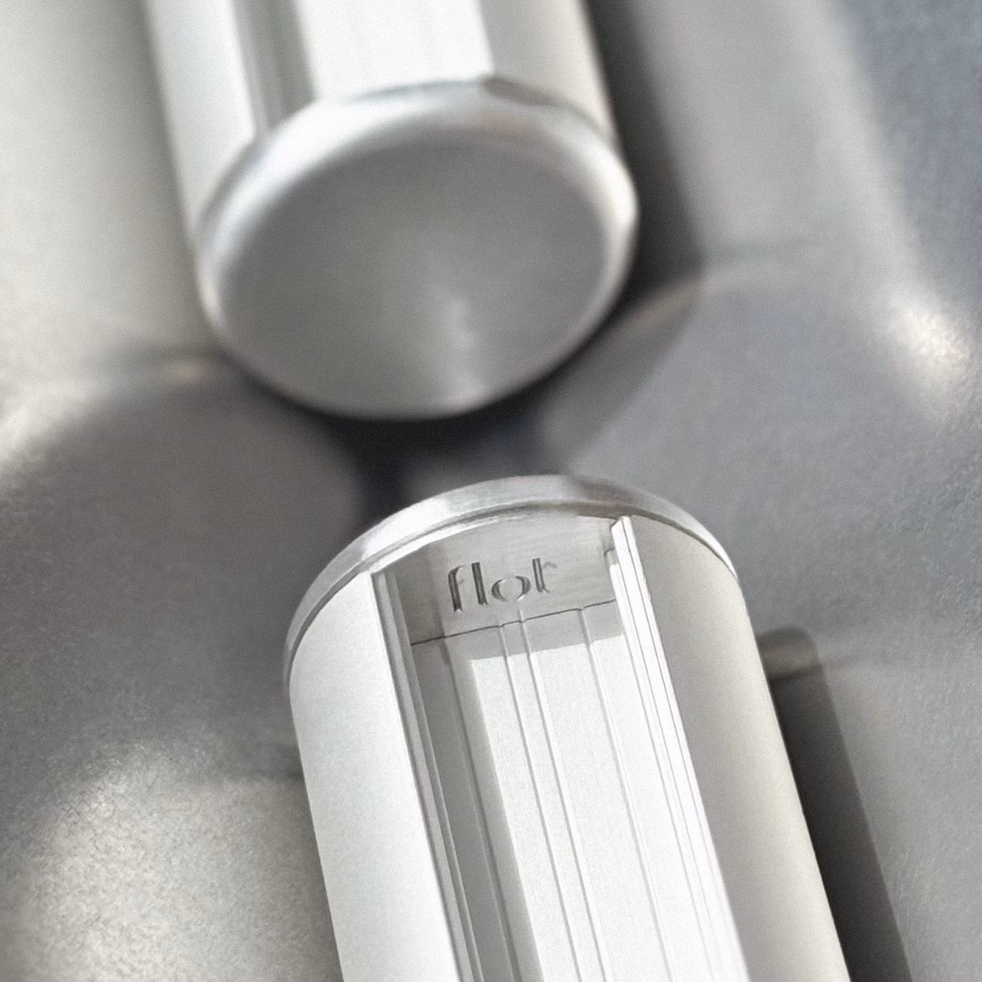 Gros plan sur un appareil cylindrique blanc avec le logo « flot » gravé, posé sur une surface métallique grise réfléchissante.