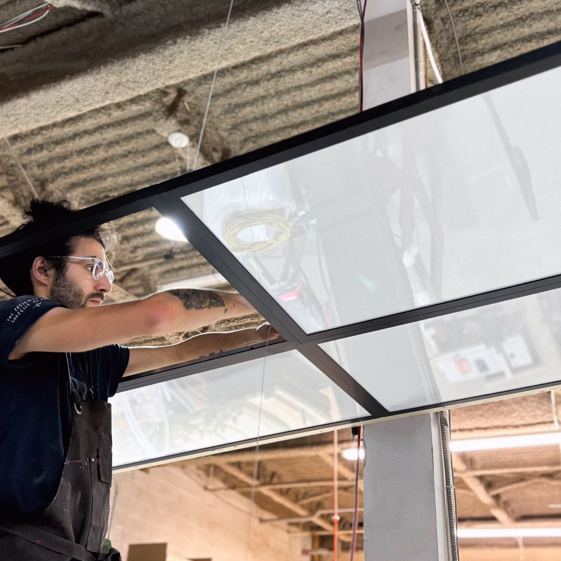 Homme avec lunette et tatouage installant un plafond suspendu lumineux dans un espace industriel.