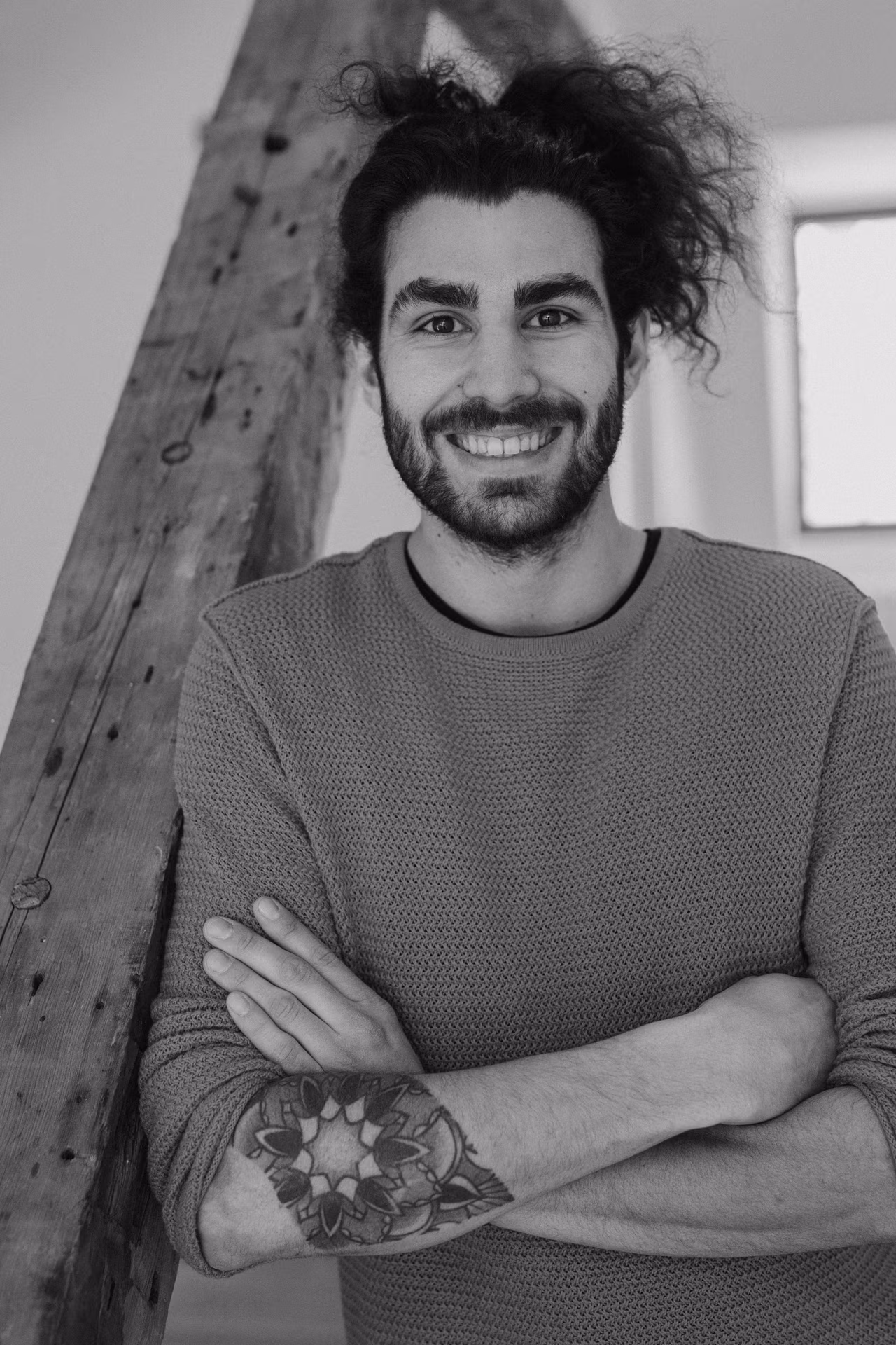 Homme souriant aux cheveux bouclés avec un pull texturé, bras croisés montrant un tatouage mandala sur son avant-bras.
