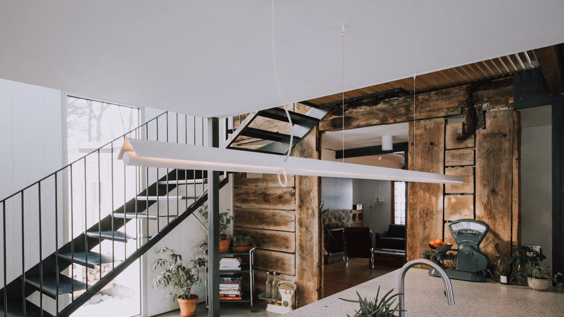 Cuisine moderne avec îlot, évier en acier inoxydable, plantes en pots, balance vintage et escalier métallique près d'un mur en bois rustique.