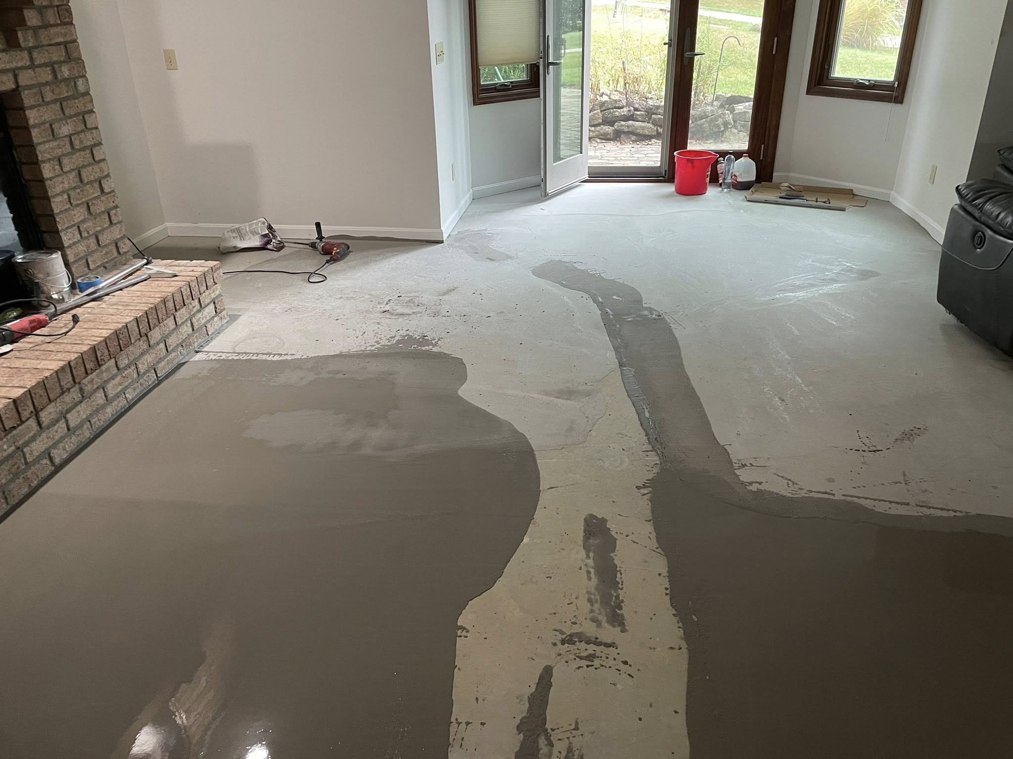 Room with partially refinished concrete floor next to a brick fireplace and glass doors leading outside.