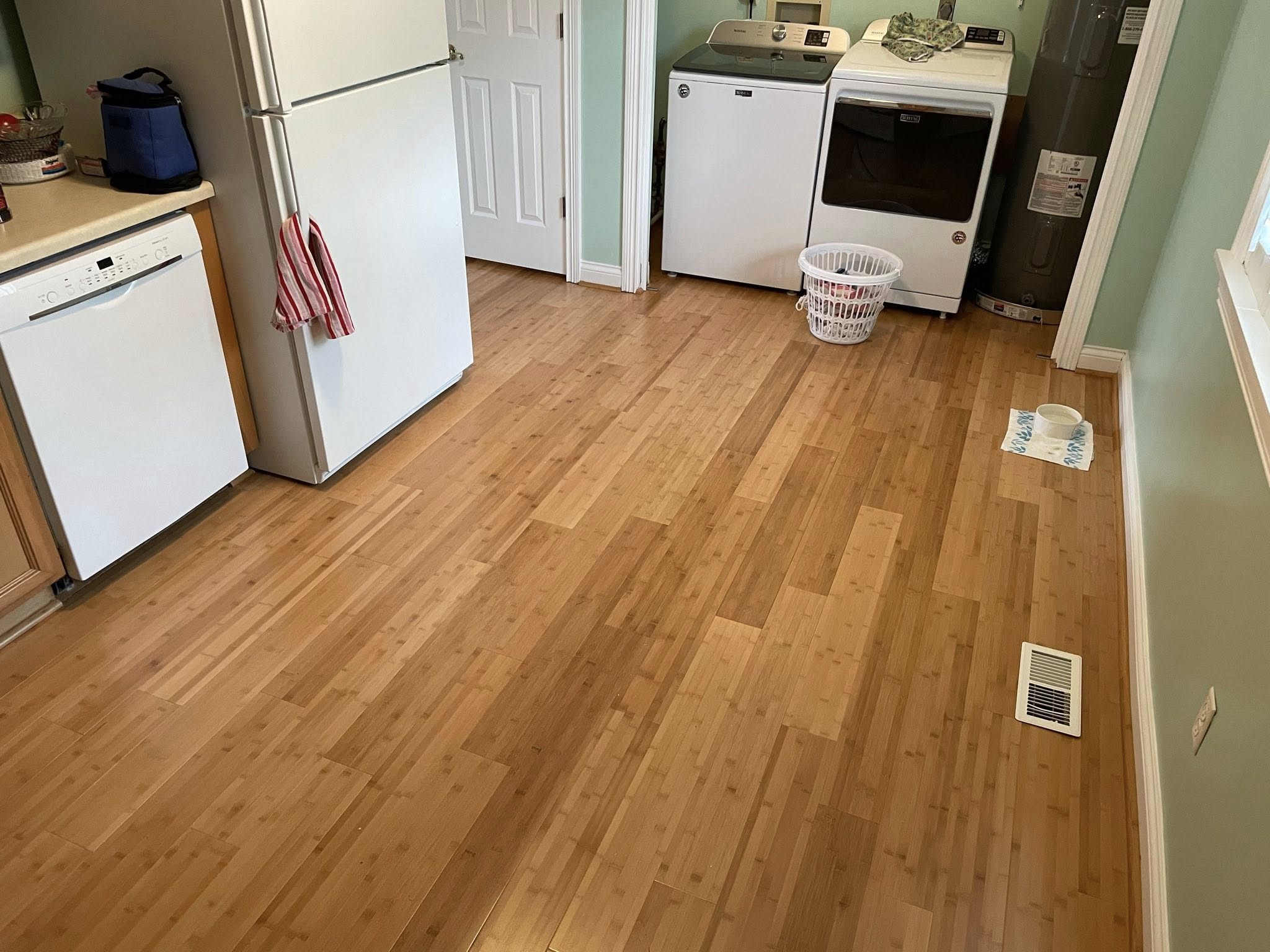 Laundry room with bamboo hardwood flooring, white washer and dryer, refrigerator, and a pet bowl on a mat next to a floor vent.