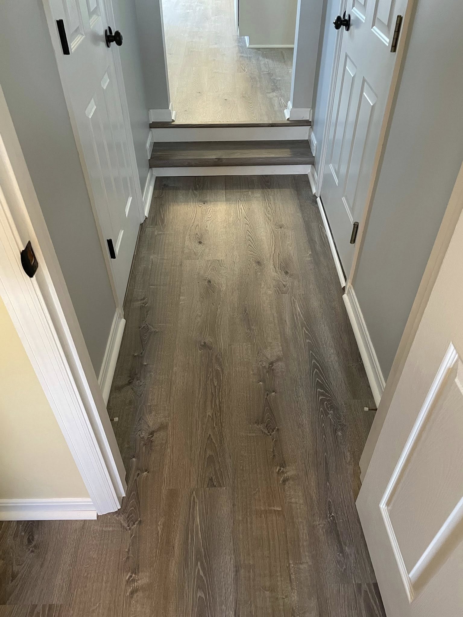 Hallway with grey walls and white doors featuring luxury vinyl plank flooring with a small step up to a lighter wood floor.