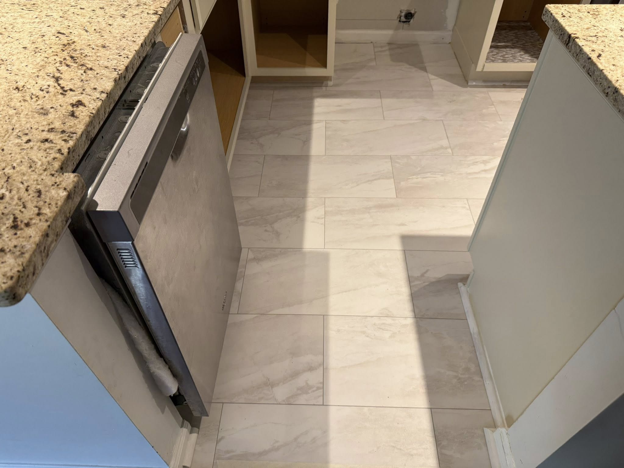 Newly installed white ceramic tile floor in a kitchen under cabinets and near a stainless steel dishwasher.