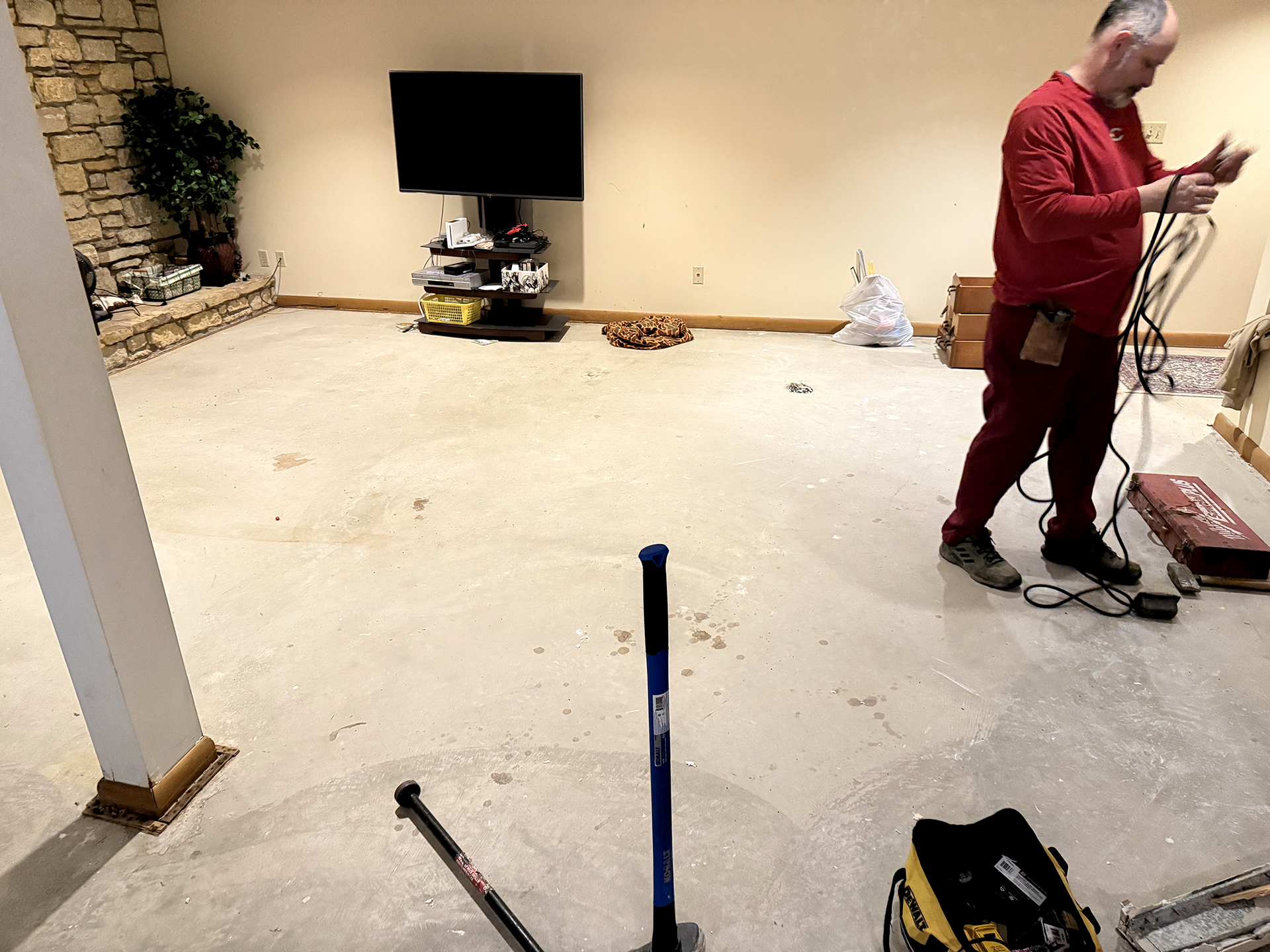 Man in red outfit working on a bare concrete floor in a room with a stone wall, TV, and scattered tools.