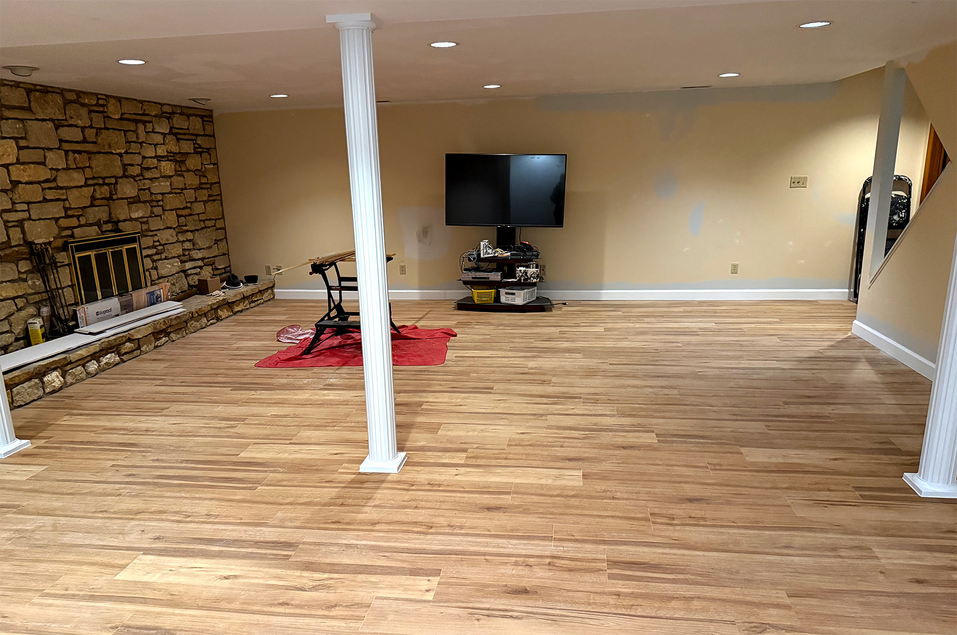 Spacious basement with wood flooring, stone fireplace on left, a mounted TV on the far wall, and two white columns in the center.