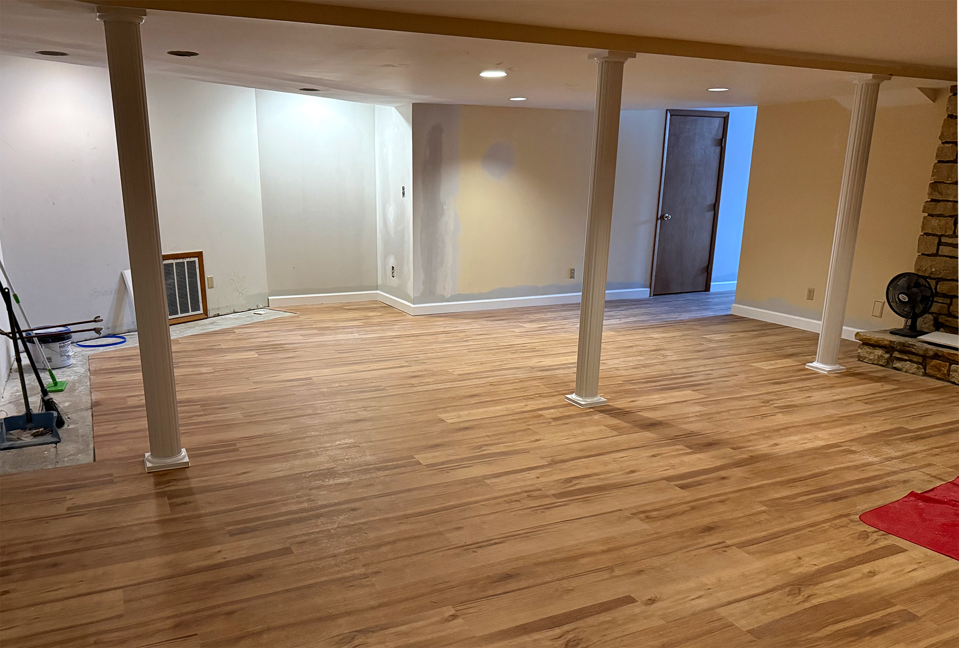Partially renovated basement with new wood flooring, white support columns, a stone fireplace, and a closed wooden door.
