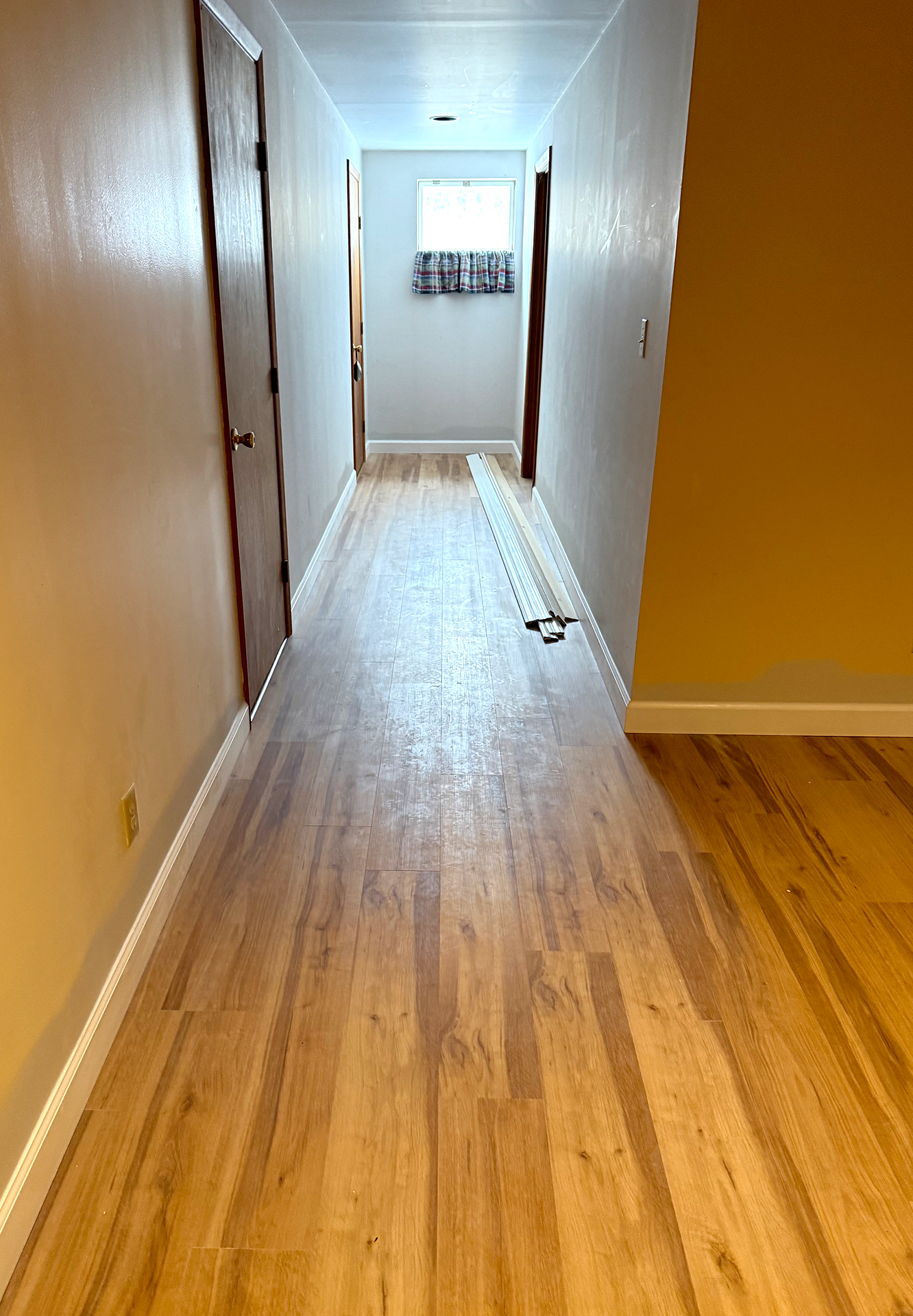 Narrow hallway with wood flooring, three closed wooden doors, a small window with a plaid curtain, and white walls.