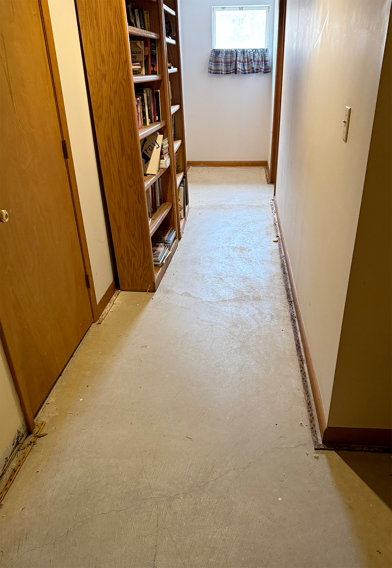 Narrow hallway with bare concrete floor, wooden doors, a bookshelf filled with books, and a small window with a plaid curtain.