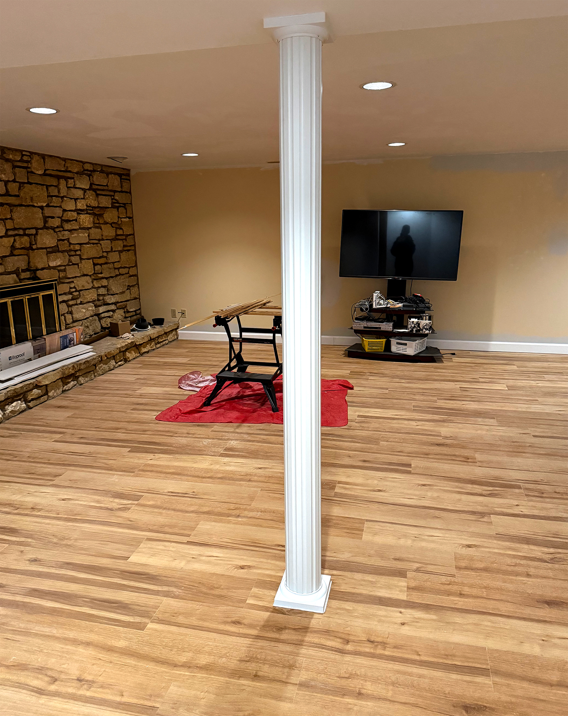 Basement room with wood flooring, a white decorative column in the center, a stone fireplace on the left, a TV on a stand against the far wall, and a workbench on a red cloth in the middle.