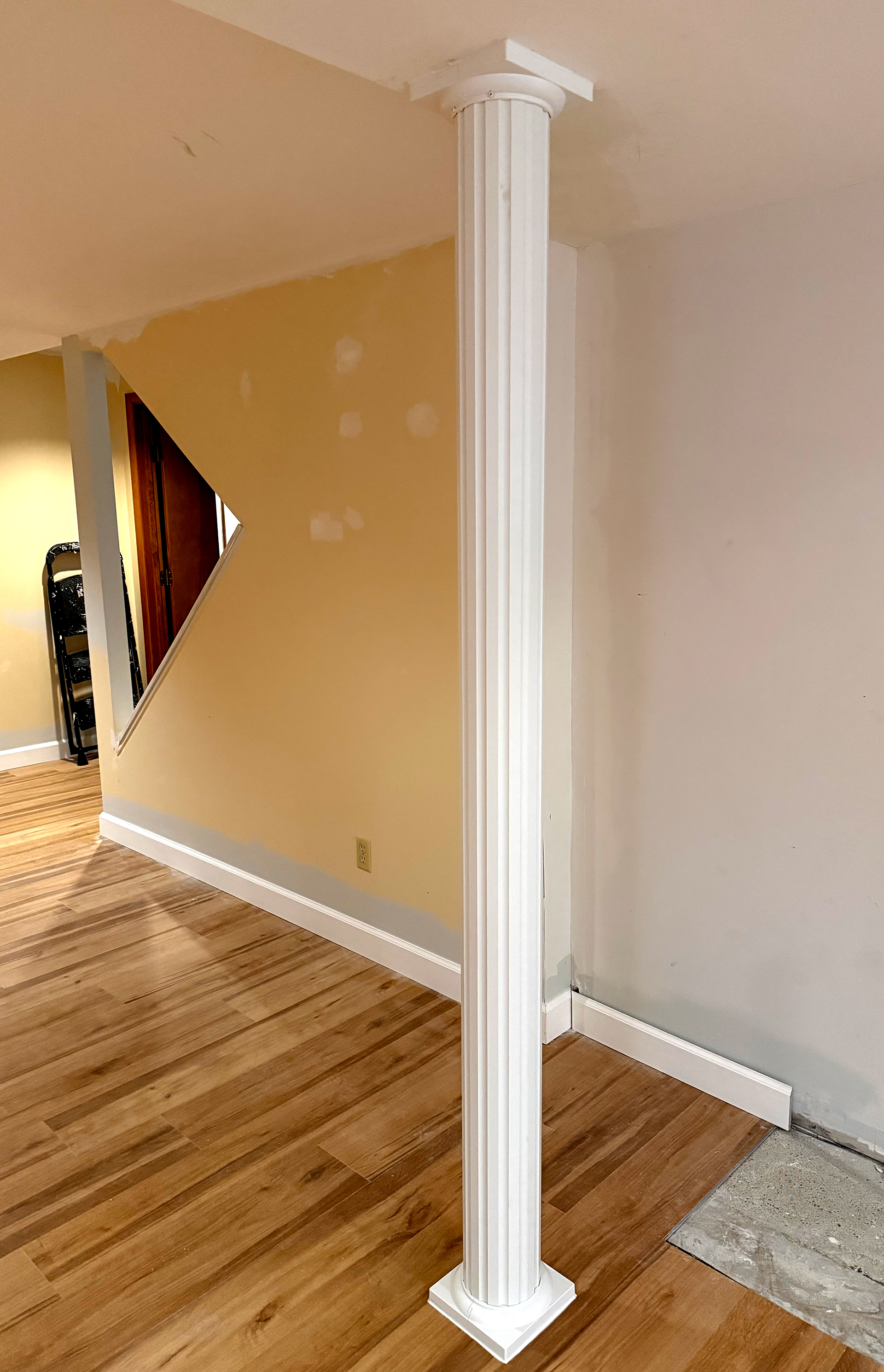 White fluted decorative column standing on wood laminate flooring near a wall with a patch of unfinished floor.