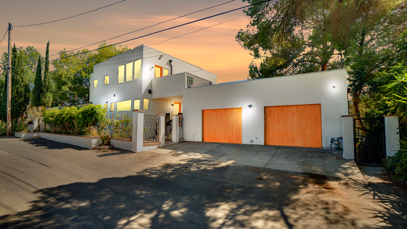 Modern two-story white house with large windows, two wooden garage doors, a gated entrance, and surrounding trees at sunset.