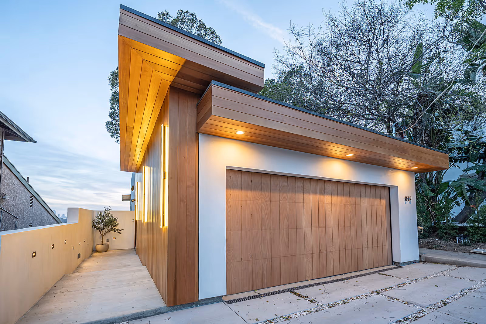 Modern house exterior with wooden garage door, angled wooden roof, illuminated wall lights, and concrete driveway.